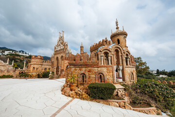 Obraz premium Castillo Monumento Colomares in Benalmadena, Malaga shows turrets, horseshoe arches, and ornate spires by a pale tiled courtyard under heavy clouds and even light.