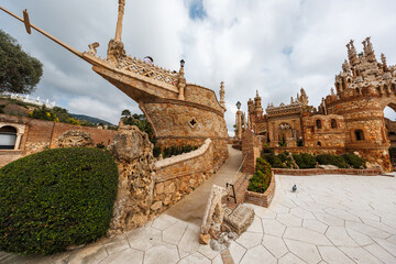 Visitors walk terraced paths at Castillo Monumento Colomares, Benalmadena, as a pigeon crosses hexagonal paving. Ornate turrets and a ship prow stand under overcast light. © True Pixel Art