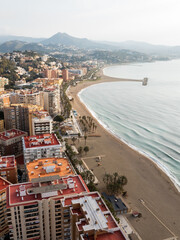 Fototapeta premium Aerial coastal view of Malaga, Spain shows La Malagueta and Pedregalejo, red roofed apartments, palm promenade, small breakwater, hazy Montes de Malaga, late afternoon light.