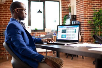 Businessman in brick wall workspace researching marketing trends on laptop. Male entrepreneur tracking company financial data on personal computer, working diligently in modern startup office.