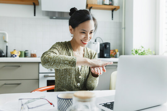 Funny good-looking deaf-mute woman using nonverbal communication to have online conversation via laptop sitting at her kitchen table, gesturing and smiling while speaking sign language