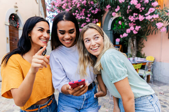 Multiracial young female friends looking at smartphone outdoors. Happy women sharing digital moments while exploring a city together. Technology and friendship concept.