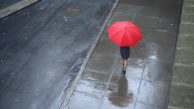 Commuter walking with red umbrella on wet city sidewalk during rain. High angle view.