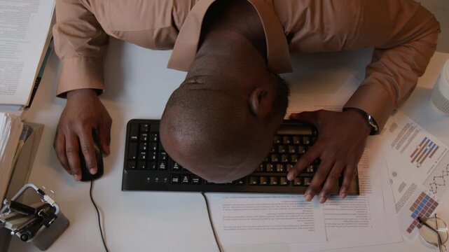 Top view shot of African American man asleep face down on keyboard at desk, holding mouse in modern office