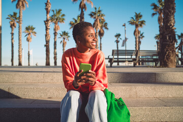 Young woman seated on city steps, holding smartphone and smiling toward the distance, reflecting...