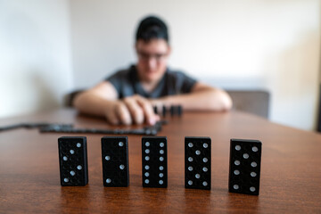 Black domino tiles stand upright in sharp focus on a table, with a blurred teenager holding a...
