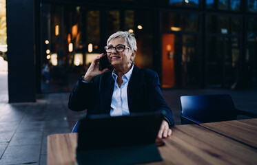 Smiling senior woman on phone call seated at table with tablet, combining personal expression with...
