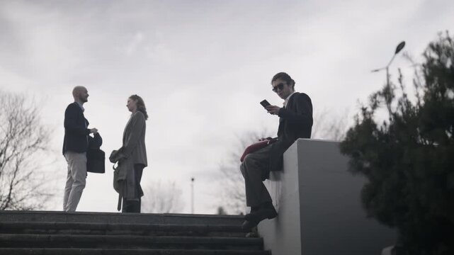 Business professionals hold an informal meeting on city steps while another person uses a smartphone. Two colleagues discuss a project outdoors, observed by a young man occupied with his phone.