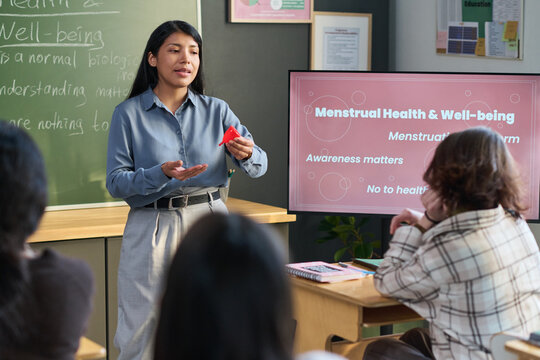 Young adult Hispanic woman standing in classroom holding menstrual cup, explaining menstrual health to group of teenagers seated at desks, educational presentation on screen