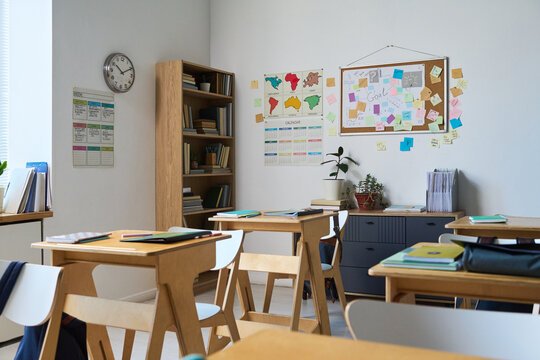 Empty classroom showing wooden desks and chairs arranged neatly, educational posters and maps on wall, bookshelves filled with books, bright daylight streaming through window, no people present