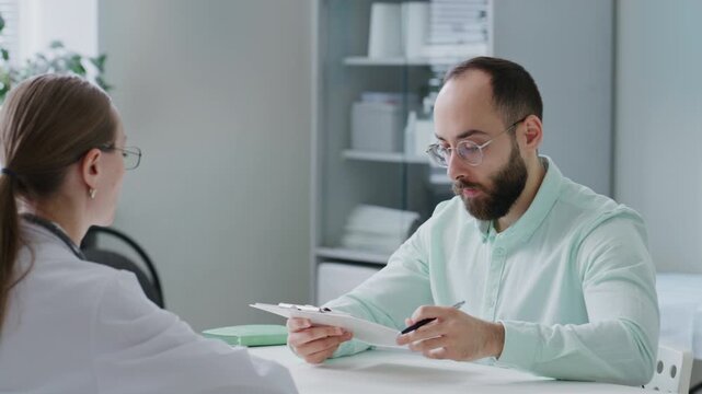 Male patient talking with female doctor and signing medical consent form during consultation at clinic, discussing treatment and completing healthcare paperwork