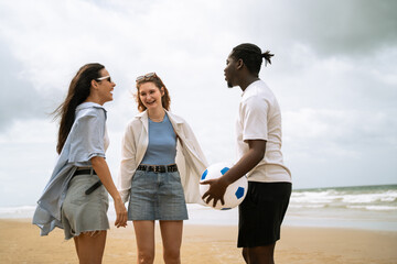 Diverse group of happy young friends laughing and talking on a sandy beach. A man holding a soccer ball stands with two women enjoying a summer vacation outdoors under a cloudy sky © winnievinzence