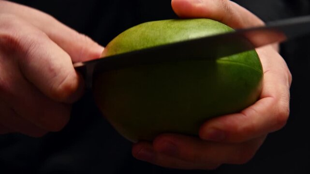 Hands slicing yellow mango fruit with knife on dark countertop surface