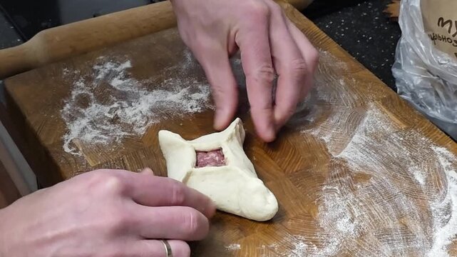 Man folds raw dough triangle with meat filling on wooden board. Cook prepares traditional pastry at home. Culinary process of making food from flour and meat.