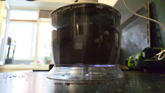 Chopper grinding dark ingredients inside transparent container. Kitchen food processor operating during meal preparation. Appliance pulverizing solid food item indoors.