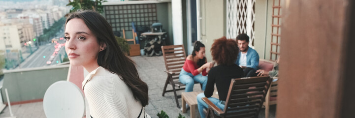 Fototapeta premium Young woman looks at the city while standing on the balcony, his friends are chatting in the background. Panoramic