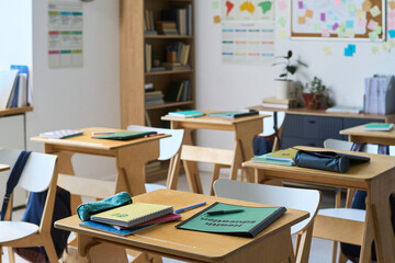 Empty classroom showing wooden desks with notebooks, folders, and stationery scattered on surfaces, bookshelves filled with books in background, educational posters and bulletin board on wall © pressmaster