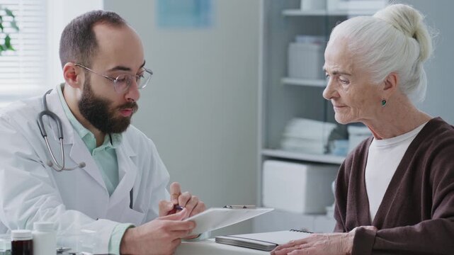 Male doctor reviewing patients records on clipboard, explaining diagnosis and treatment plan to elderly woman during medical appointment in clinic