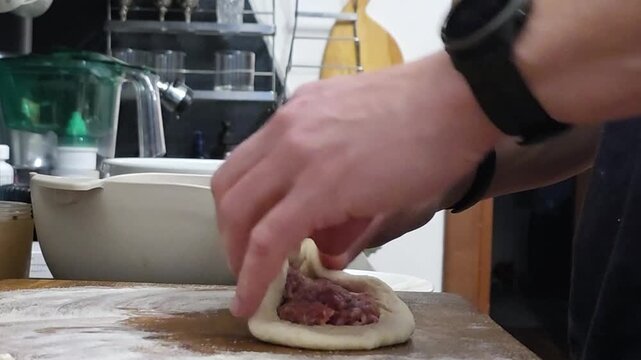Man preparing dough with meat stuffing on wooden table. Cook folding pastry for dumpling making at home. Culinary skill, food preparation and homemade meal craft.