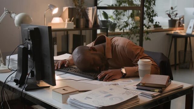 Tired from overworking African American man sleeping face down on computer keyboard at desk in modern office