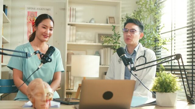 Asian medical doctor hosting a healthcare podcast interview with a nurse inside a bright clinic office. Professional consultation recording with microphones and laptop discussing treatment, patient	