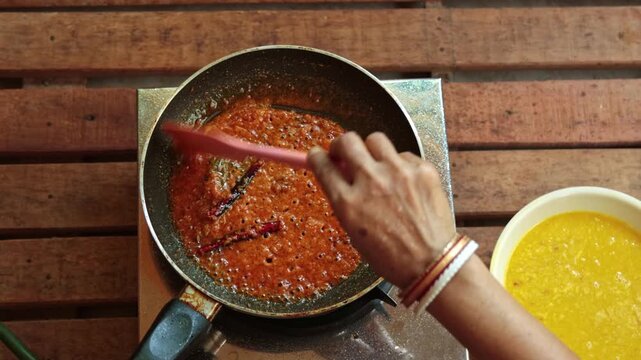 Top-down close-up of a woman&rsquo;s hand adding a spicy tempering (tadka) of red chilies, cumin seeds, and curry leaves into a pan of yellow dal lentils, sizzling in hot ghee on a wooden kitchen counter.