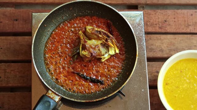 Top-down view of fried fish heads being added with tongs to a vibrant, simmering red masala gravy in a black frying pan. Traditional Indian cooking process on a portable stove, featuring dried red chi