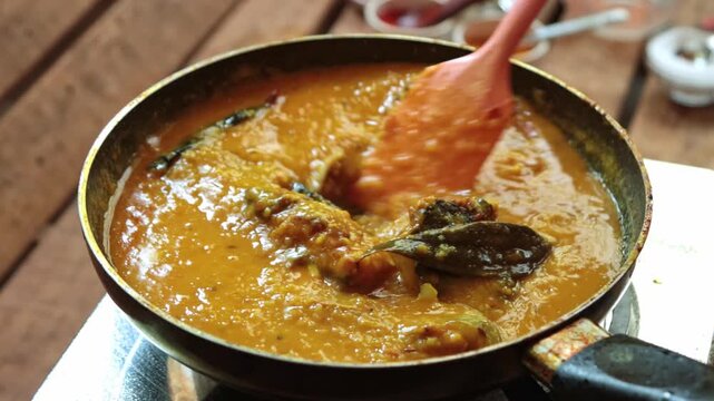 Close-up of vibrant, spicy fish curry bubbling and being stirred in a pan. Features golden gravy, fried fish pieces, and green chilies. Ideal for culinary, travel, and South Asian food content.