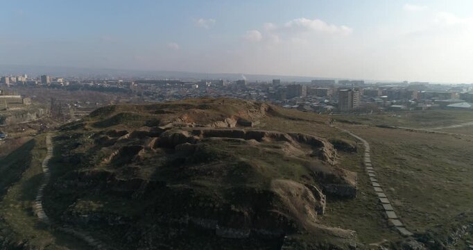 Breathtaking aerial shot of the ancient Erebuni Fortress ruins situated on a prominent hill. The drone flies forward revealing the archaeological site with the vast cityscape of Yerevan in the backgro