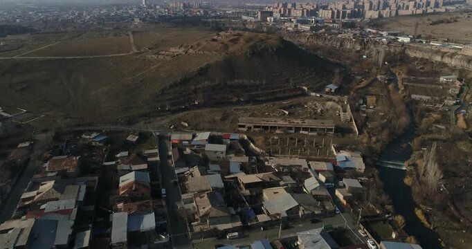 Breathtaking aerial shot of the ancient Erebuni Fortress ruins situated on a prominent hill. The drone flies forward revealing the archaeological site with the vast cityscape of Yerevan in the backgro
