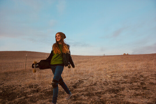 Young woman in jeans Wearing Kneepads walking in a rural landscape Carrying a Longboard, Utah, USA