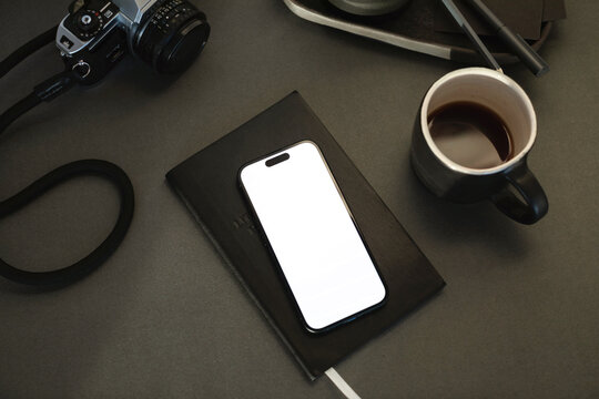 Close-up of a mobile phone with a blank screen on a notebook with a cup of black coffee and camera on a desk