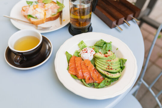 Close-up of a salmon, avocado, and poached egg salad, shrimp toast with a poached egg and greens and a herbal tea on a table at an outdoor cafe