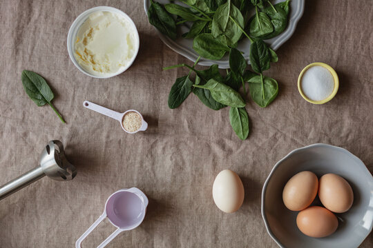 Overhead view of healthy gluten free pancake ingredients and kitchen utensils including eggs, fresh spinach, mascarpone cheese, salt, and psyllium on a linen tablecloth