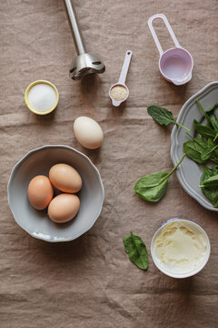 Overhead view of healthy gluten free pancake ingredients and kitchen utensils including eggs, fresh spinach, mascarpone cheese, salt, and psyllium on a linen tablecloth