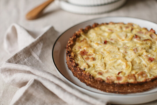 Close-up of a homemade savoury gluten free keto onion and bacon tart with an almond flour crust on a plate with a stack of plates, a cake server and napkins