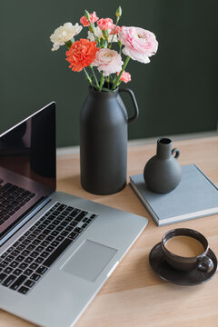 Laptop computer with a blank screen, a cup of coffee, a bunch of pink and orange carnation and ranunculus flowers in a ceramic vase and notebooks