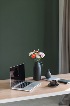 Laptop computer with a blank screen, a cup of coffee, a bunch of pink and orange carnation and ranunculus flowers in a ceramic vase and notebooks