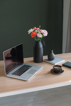 Laptop computer with a blank screen, a cup of coffee, a bunch of pink and orange carnation and ranunculus flowers in a ceramic vase and pens in a glass on a desk