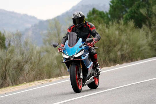 Motociclista con equipamiento completo pilotando una motocicleta deportiva azul con llantas rojas en una carretera de monta&ntilde;a.