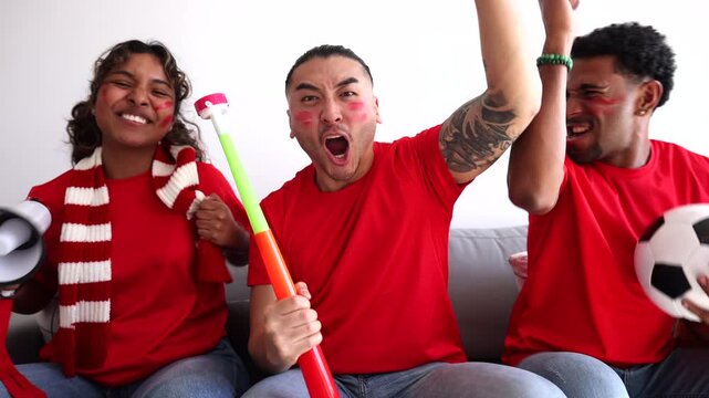 Three diverse friends, wearing red team jerseys and face paint, celebrating a goal with excitement, screaming and cheering while watching a soccer match at home on a white background