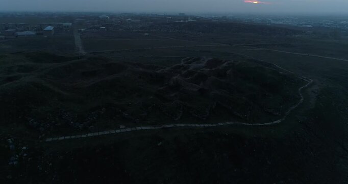  Aerial drone video reveals an ancient archaeological site on a hill at sunset. The camera tilts down to show excavated walls and trenches, with a distant city in the background.