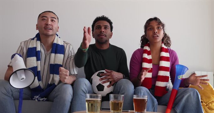Three multiethnic friends watching a football match on television with opposing team scarfs, experiencing a rollercoaster of emotions from tension and disappointment to celebration and mockery