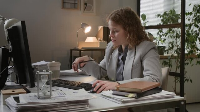 Woman in business suit intently working at computer desk while tracking time with hourglass in office