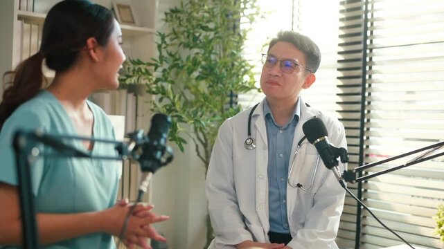 Asian medical doctor hosting a healthcare podcast interview with a nurse inside a bright clinic office. Professional consultation recording with microphones and laptop discussing treatment, patient	