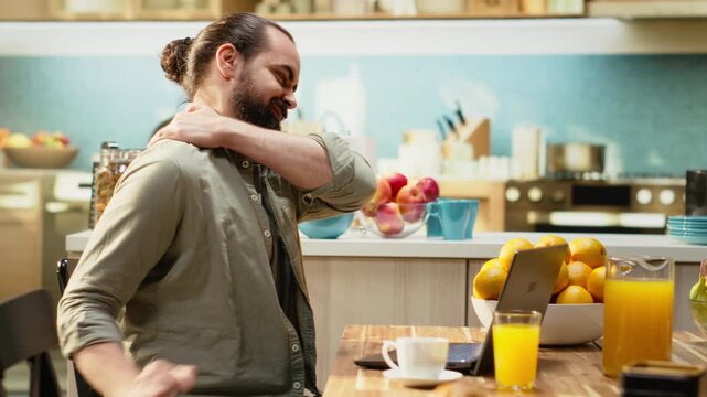 Man with neck strain does self massage to relieve pain and pressure, stiffness and physical fatigue due to incorrect chair posture. Freelancer stretching at home, teleworking in the kitchen.