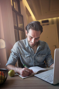 Man smiling, writing notes in a notebook while working on a laptop at home, focusing on productivity, inspiration, and distance learning concepts