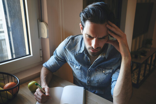 Concerned man feeling stressed and having headache while thinking and writing notes in a notebook, managing work or personal challenges at a home working desk