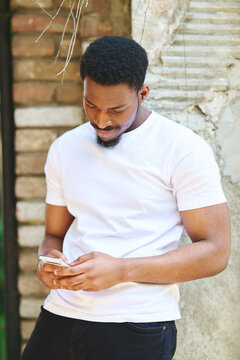 Young black man in a white t-shirt actively communicating using a mobile phone, focused on his smartphone screen, connecting and networking in an urban environment