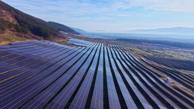 Rows of solar panels stretch across a field under a blue sky with mountains visible in the distance. The sunlight reflects off the panels, showcasing renewable energy production.